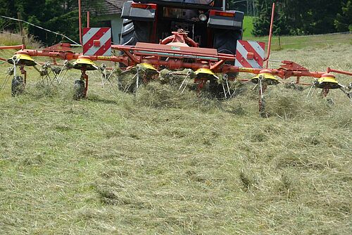 Sur la photo, on peut voir un secoueur de foin attelé à un tracteur, et au premier plan, du foin séché dans une prairie. 