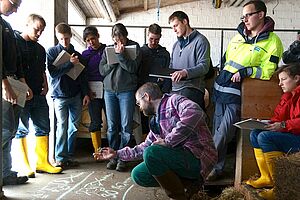 Un groupe de jeunes se tient en cercle dans une étable autour d'un professeur qui écrit une formule à la craie sur le sol.er mit Kreide eine Formel auf den Boden schreibt. 
