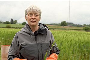 Une femme en tenue de travail se tient dans un champ de céréales et tient un épi dans sa main.